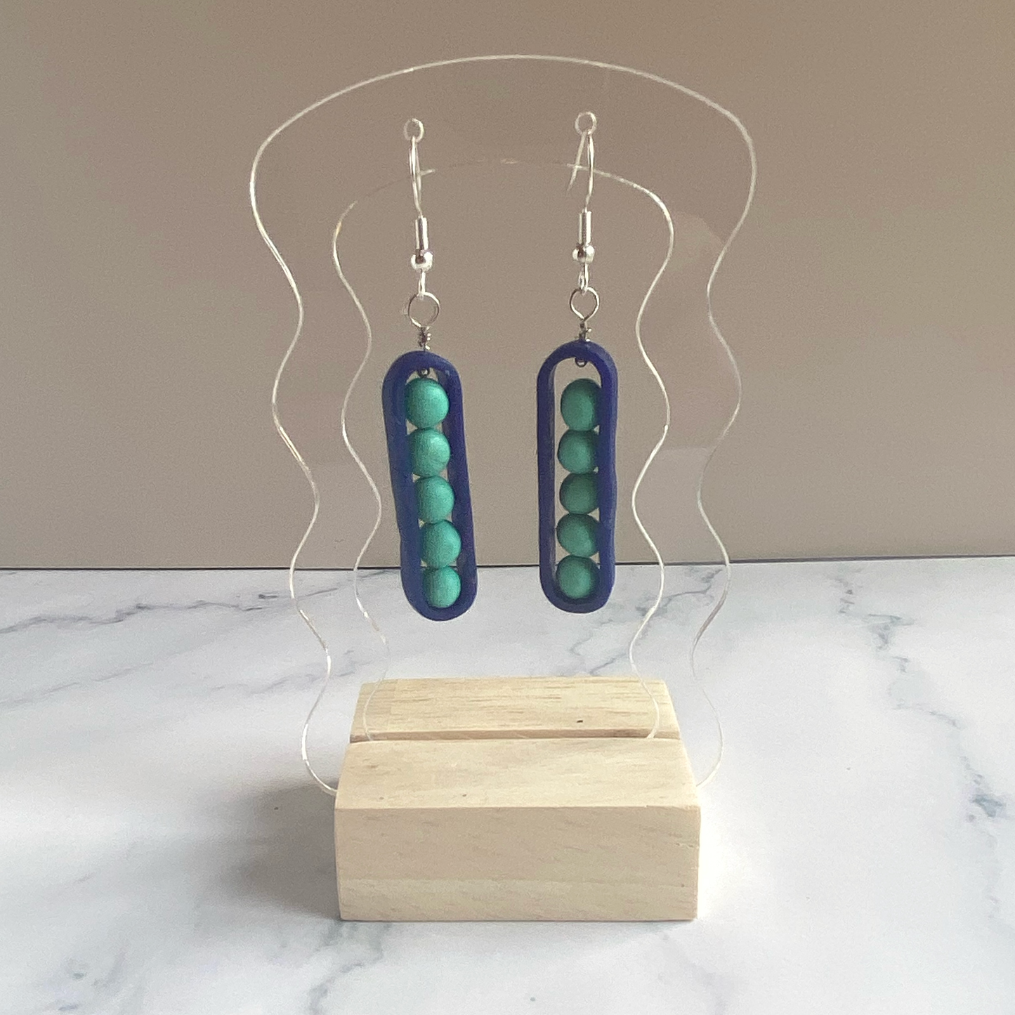 Earrings with aqua 'peas' in royal blue 'peapos' on a clear acrylic stand with a beige background