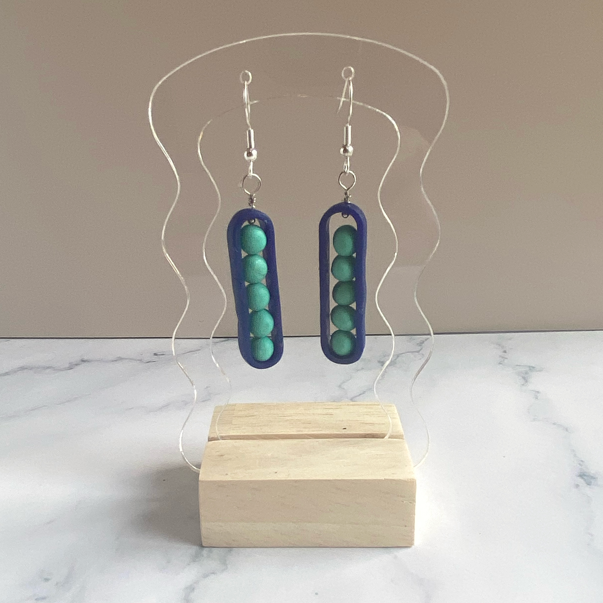 Earrings with aqua 'peas' in royal blue 'peapos' on a clear acrylic stand with a beige background
