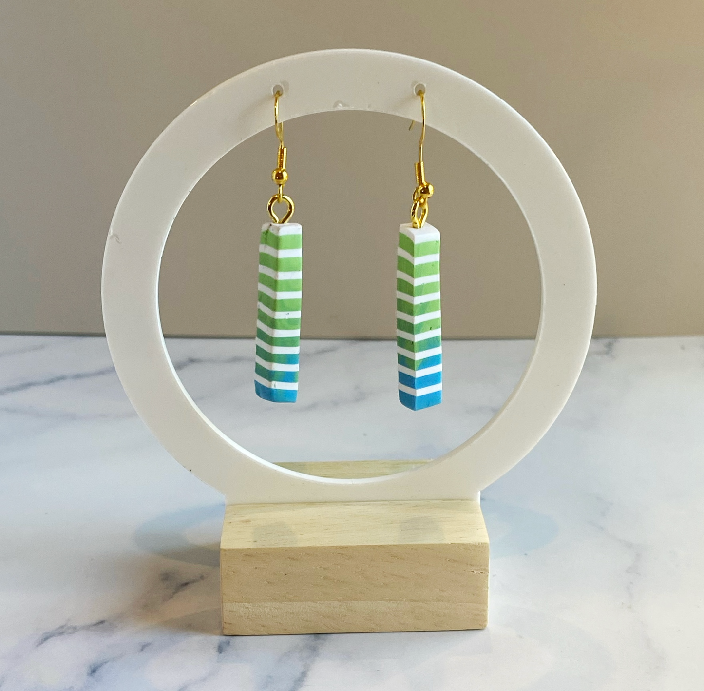 Green and blue striped earrings on a white circular stand with a wooden base.