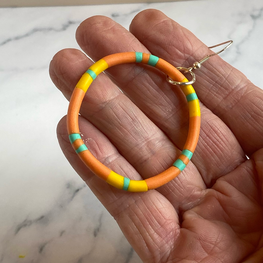 Orange hoop earrings with turquoise and yellow accents held in a hand against a marble background